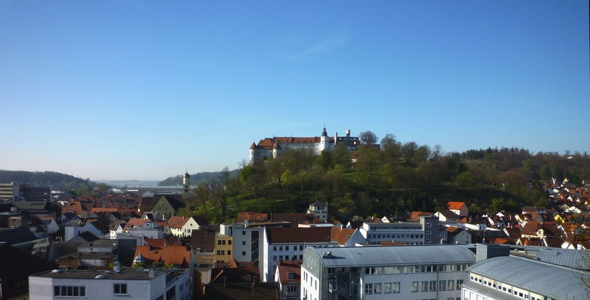 Blick über die Dächer von Heidenheim an der Brenz auf den bewaldeten Schlossberg mit Schloss Hellenstein unter klarem blauem Himmel.