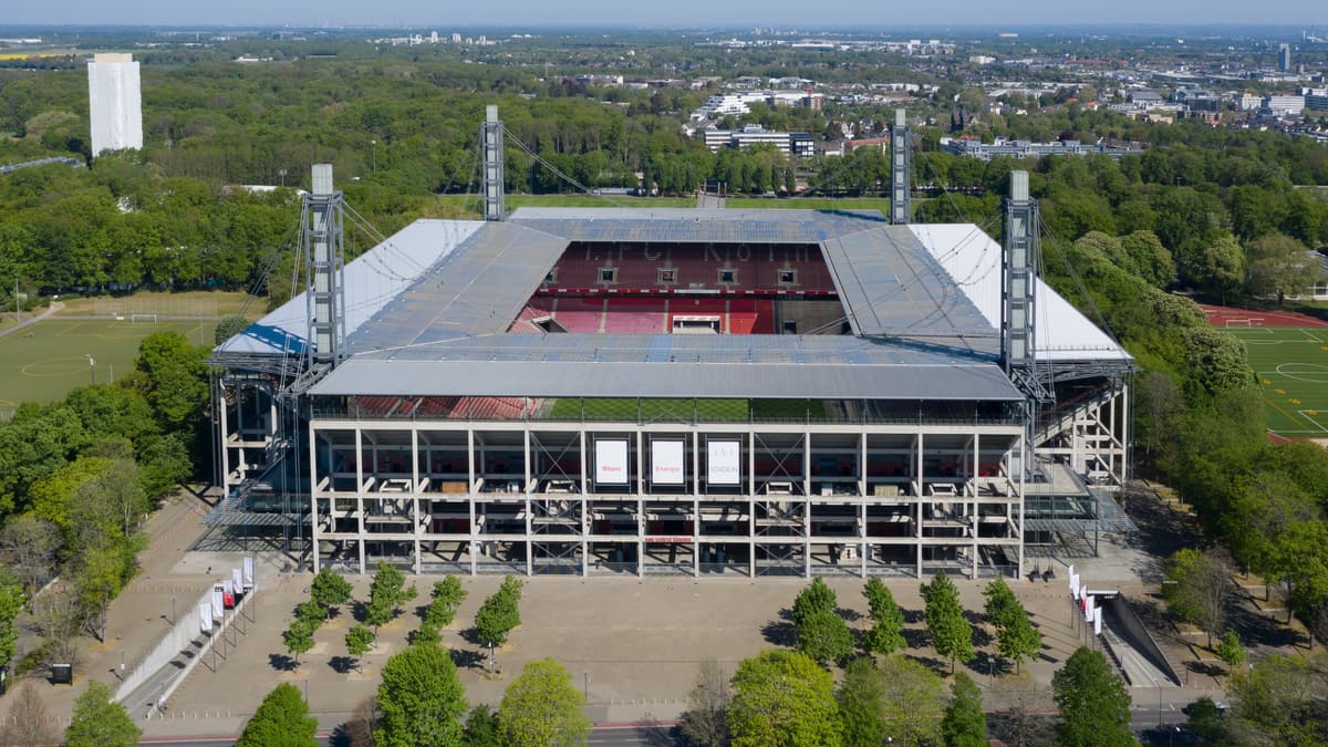 Luftaufnahme des RheinEnergieStadions mit seinen vier Pylonen, umgeben von Bäumen und Sportanlagen in Köln-Müngersdorf.