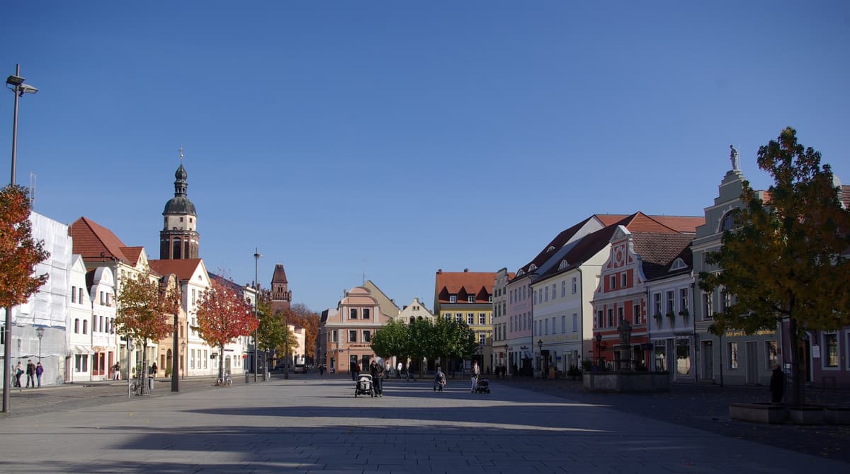 Der weite Altmarkt in Cottbus mit Bürgerhäusern und herbstlichen Bäumen, im Hintergrund der Turm der Oberkirche St. Nikolai unter tiefblauem Himmel.