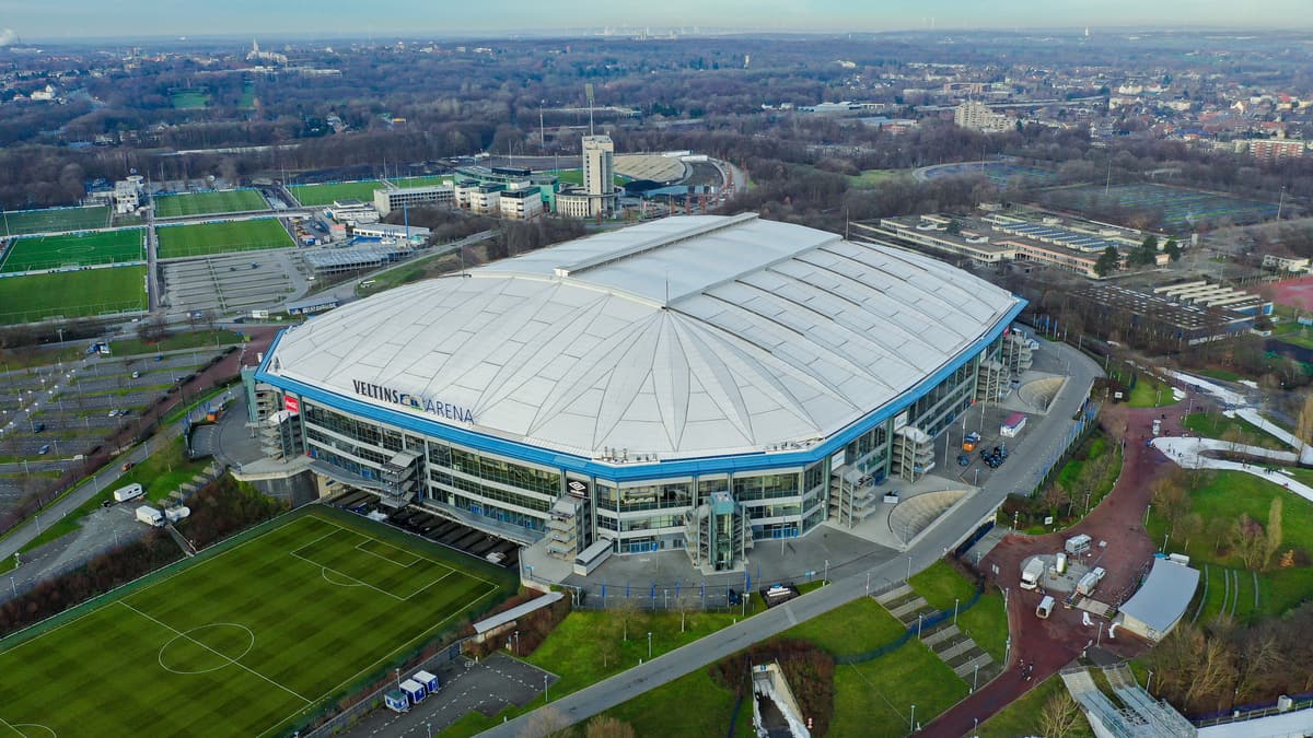 Luftbild der Veltins-Arena mit weißem Dach und blauer Fassade, daneben Trainingsplätze.