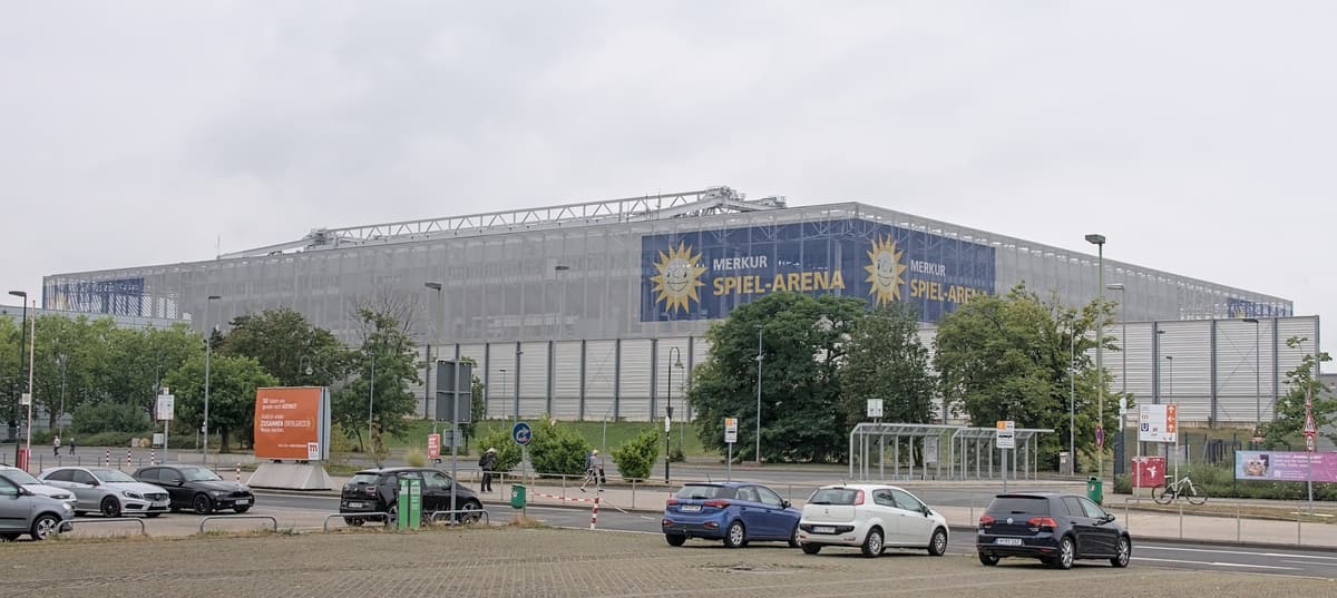 Die Merkur Spiel-Arena mit Schriftzug an der Fassade hinter einer Straße und einem Parkplatz.