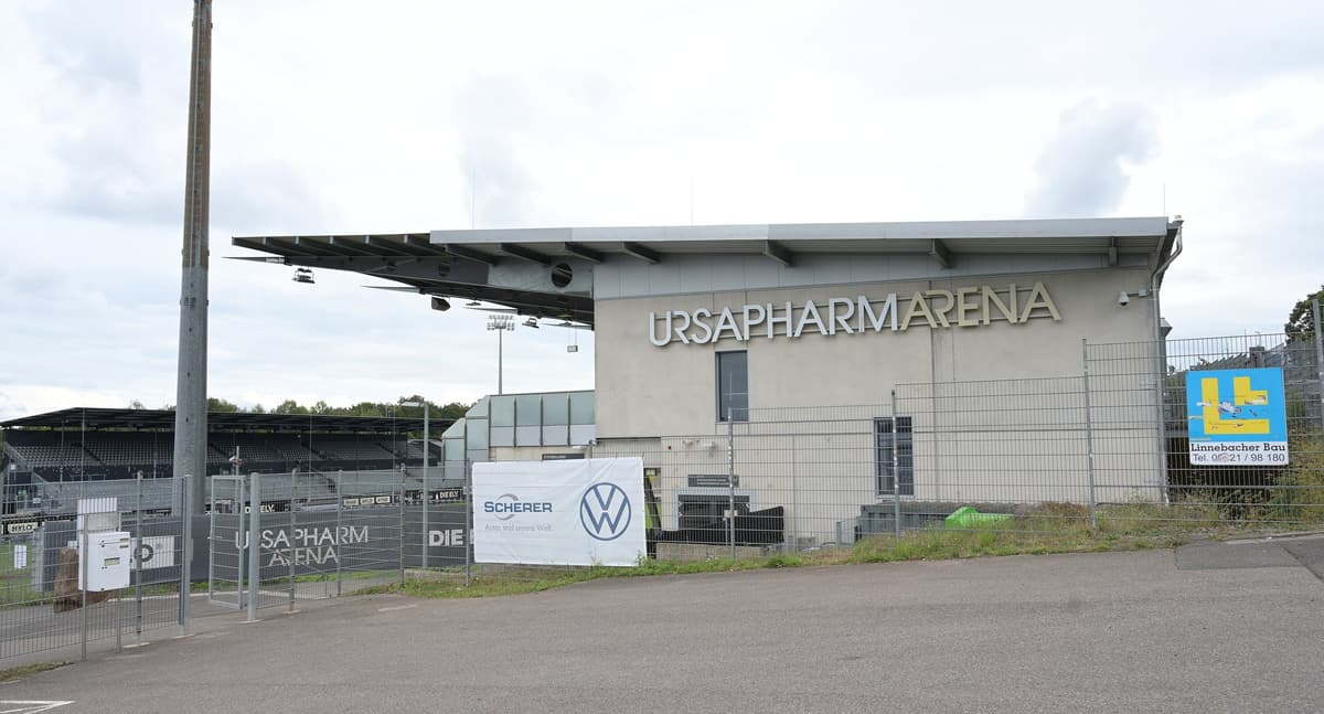 Außenansicht der Ursapharm-Arena an der Kaiserlinde: Tribünengebäude mit großem Schriftzug „Ursapharm Arena“ hinter einem Bauzaun, davor ein leerer Parkplatz.