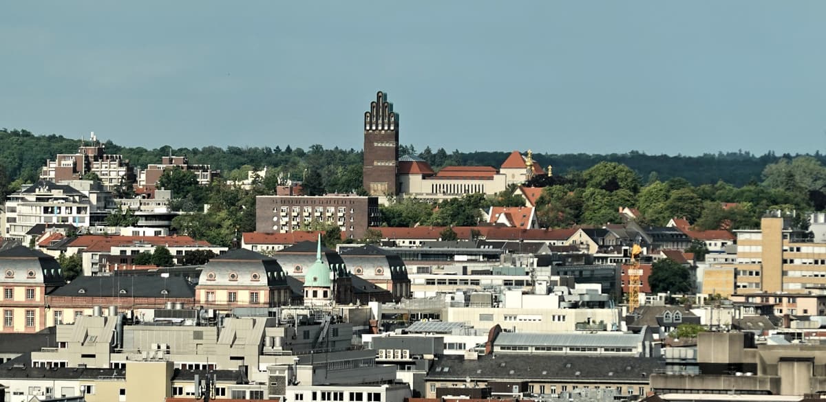 Blick über die Innenstadt von Darmstadt auf die Mathildenhöhe mit dem Hochzeitsturm vor bewaldeten Hügeln.